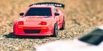 To små blå BTD Drifter fjernstyrede biler i fuld fart på en detaljeret miniature racerbane med dækstakke.