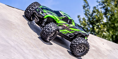 Sort Traxxas Mini Maxx monster truck med blå detaljer kører hurtigt gennem mudret terræn.