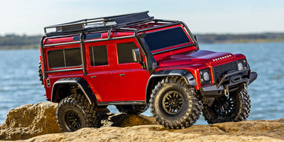 Mørkerød Traxxas TRX-4 Defender crawler kører gennem mudret terræn i naturen.